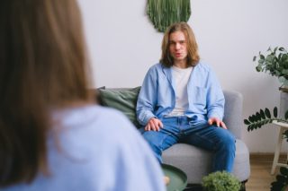 Concerned male client siting on sofa while having conversation about problems with faceless psychologist during psychotherapy appointment in light office