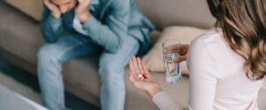woman holding glass of water and pills while man suffering from headache