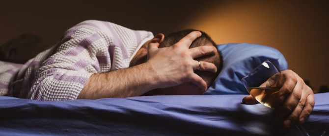 A man with a headache holds a glass of wine in bed